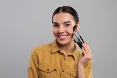 Farklı makyaj fırçaları olan mutlu kadın açık gri arka planda