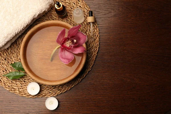 Water with beautiful flower in bowl, bottles of essential oil and candles on wooden table, flat lay with space for text. Aromatherapy treatment