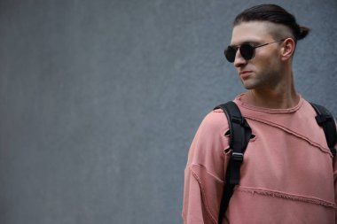 Gri duvarın yanında şık güneş gözlüklü ve sırt çantalı yakışıklı genç adam, mesaj için yer var.