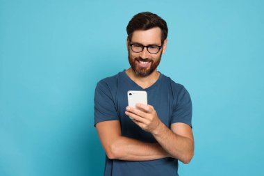 Açık mavi arka planda akıllı telefona bakan mutlu bir adam.