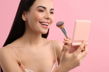 Kozmetik cep aynalı güzel bir kadın pembe arka plana makyaj yapıyor.