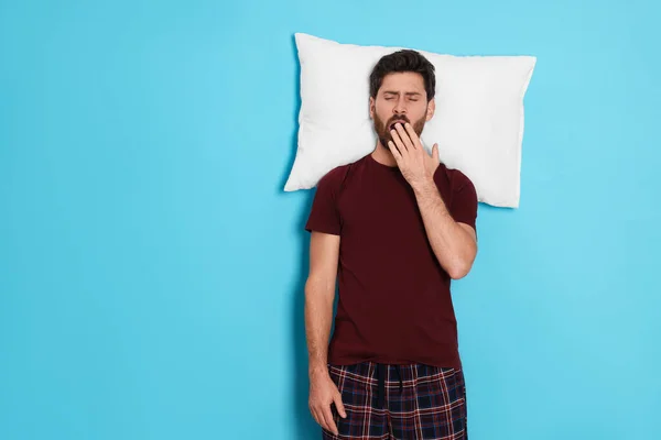 Tired man with pillow yawns on light blue background, space for text