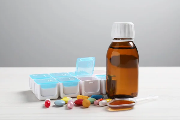 Bottle of syrup, dosing spoon and pills on white table against light grey background. Cold medicine