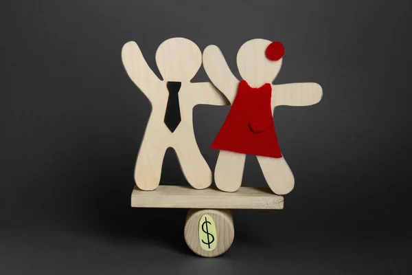 Gender pay gap. Wooden figures of man and woman on miniature seesaw against black background