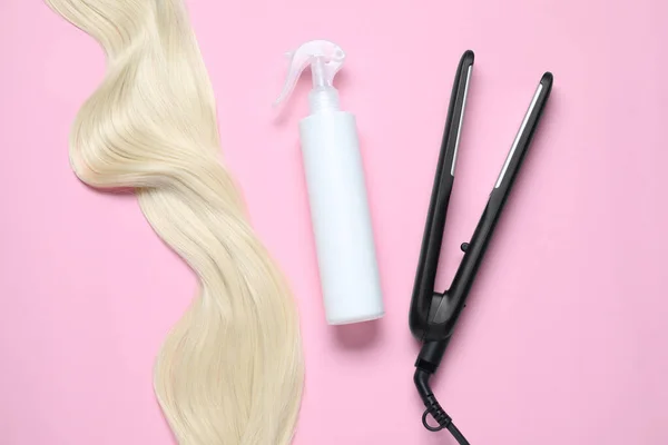 Spray bottle with thermal protection, iron and lock of blonde hair on pink background, flat lay