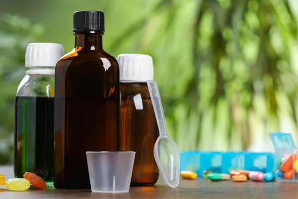 Bottles of syrup, measuring cup, dosing spoon and pills on wooden table against blurred background, space for text. Cold medicine