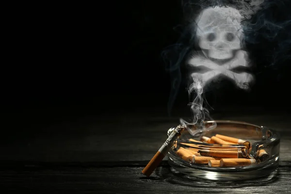 No Smoking. Skull and crossbones symbol of smoke over ashtray with stubs and smoldering cigarette on dark wooden table against black background. Space for text