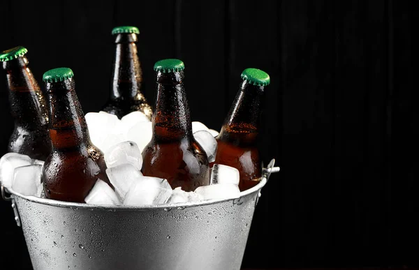 Metal bucket with bottles of beer and ice cubes on dark wooden background, closeup