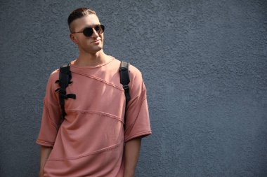 Gri duvarın yanında şık güneş gözlüklü ve sırt çantalı yakışıklı genç adam, mesaj için yer var.