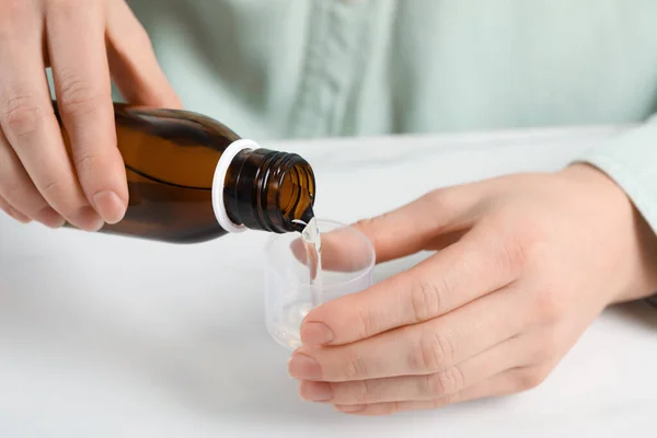 Woman pouring syrup from bottle into measuring cup at white table, closeup. Cold medicine