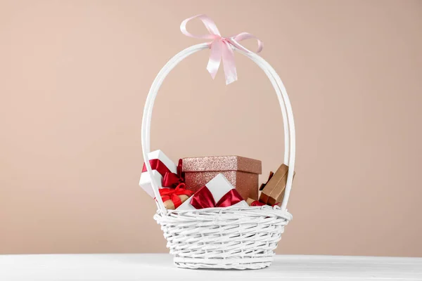 Wicker basket full of gift boxes on white wooden table against beige background