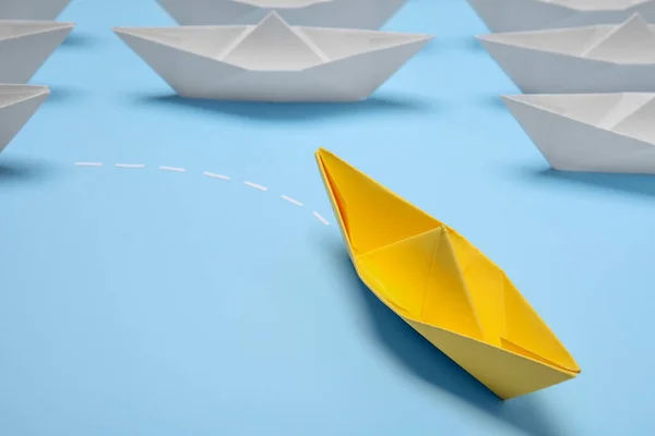 Yellow paper boat floating away from others on light blue background, closeup with space for text. Uniqueness concept