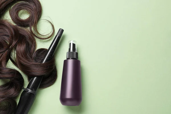 Spray bottle with thermal protection, lock of brown hair and stylish curling iron on pale green background, flat lay. Space for text