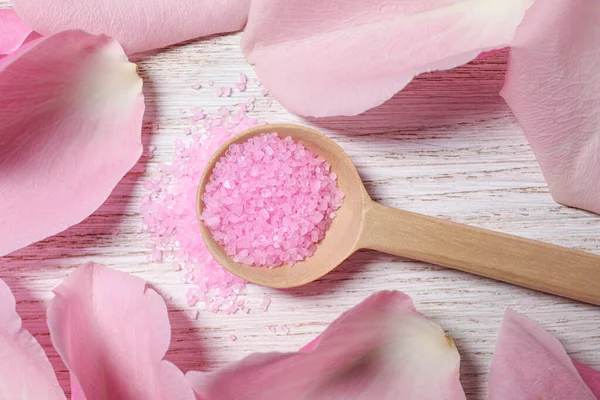 Spoon with pink sea salt and petals of roses on white wooden table, flat lay