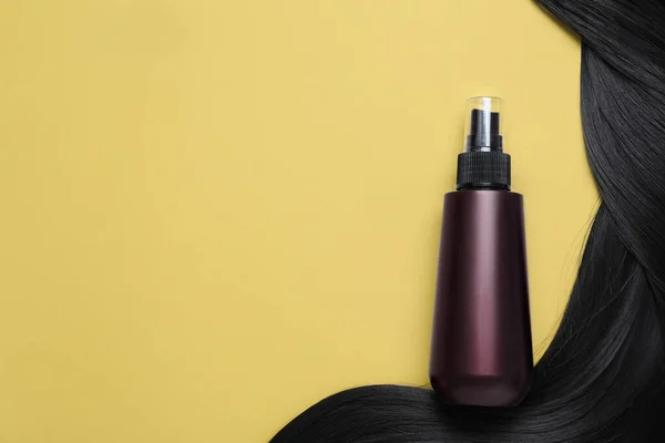 Spray bottle with thermal protection and lock of black hair on yellow background, flat lay. Space for text