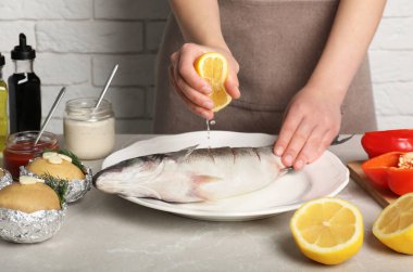 Açık gri mermer masada taze levrek balığına limon suyu sıkan kadın, yakın plan.