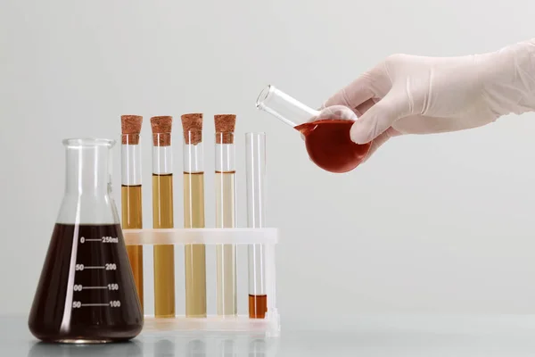 Scientist pouring brown liquid from round bottom flask into test tube on light background, closeup