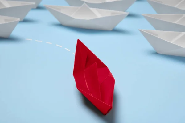 Red paper boat floating away from others on light blue background, closeup. Uniqueness concept