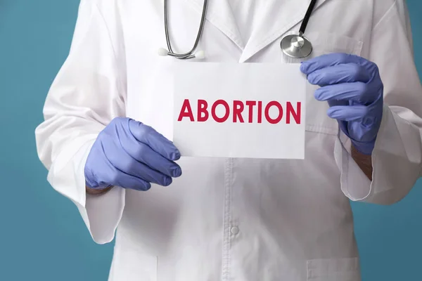 Doctor holding paper card with word Abortion on light blue background, closeup