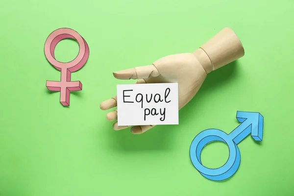 Equal pay. Wooden mannequin hand, paper note, gender symbols on green background, flat lay