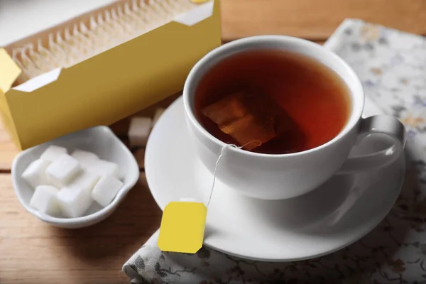 Tea bags package, sugar and cup of hot drink on wooden table, closeup
