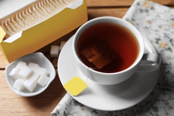 Tea bags package, sugar and cup of hot drink on wooden table, closeup