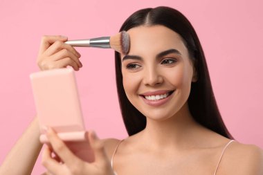 Kozmetik cep aynalı güzel bir kadın pembe arka plana makyaj yapıyor.