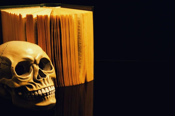 Human skull and old book on mirror table against black background. Space for text