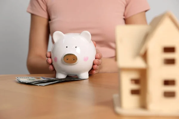 Woman holding piggy bank, house model and banknotes at wooden table, closeup