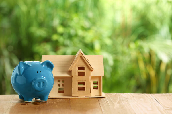 Свинка банка, маленькая модель дома и монеты на деревянном столе на открытом воздухе. Пространство для текста