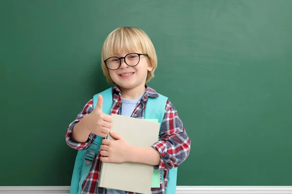 Happy little school child with notebooks showing thumbs up near chalkboard. Space for text