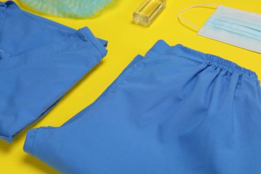 Medical uniform, face mask and antiseptic on yellow background, closeup view