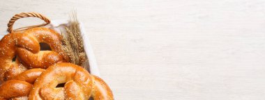 Basket with delicious pretzels and wheat spikes on white wooden table, top view with space for text. Banner design