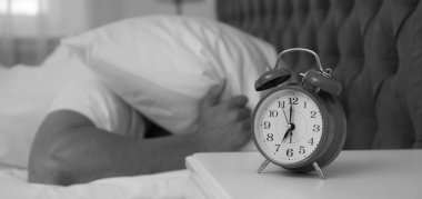 Alarm clock and man covering head with pillow in bedroom, selective focus. Black and white photography