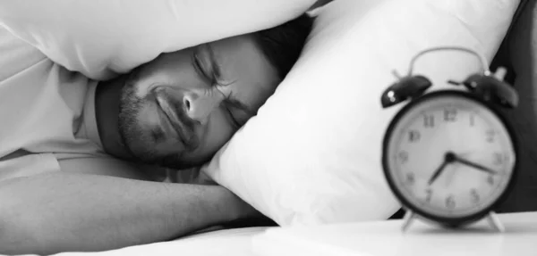 Emotional man covering ears with pillows at home in morning, selective focus. Black and white photography