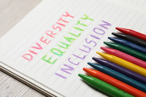 Notebook with words Diversity, Equality, Inclusion and wax pencils on wooden table, closeup