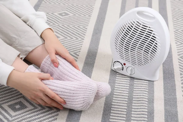 Woman warming feet near electric fan heater at home, closeup