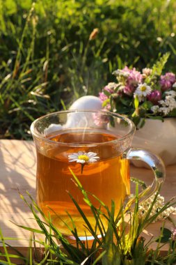 Bir fincan aromatik bitkisel çay, tokmak ve seramik havan, yeşil çimlerin üzerinde farklı kır çiçekleri.