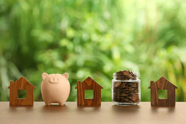 Piggy bank, jar of coins and house models on wooden table outdoors