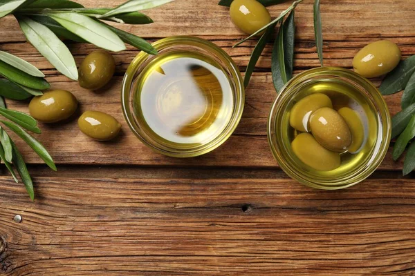 Bowls with cooking oil, green leaves and olives on wooden table, flat lay. Space for text