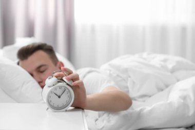 Sleepy man turning off alarm clock at home in morning, focus on hand. Space for text