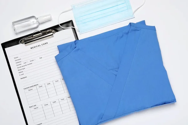 Medical uniform, face mask and clipboard on white background, flat lay
