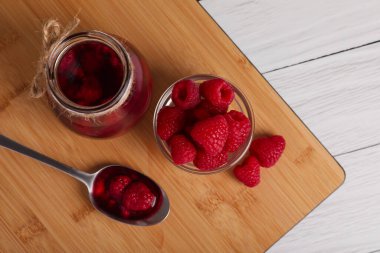 Jar of tasty canned raspberry jam and fresh berries in glass bowl on white wooden table, flat lay