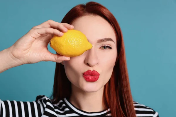 Beautiful redhead woman covering eye with lemon and blowing kiss on light blue background