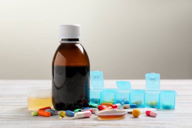 Bottle of syrup, measuring cup, dosing spoon and pills on white wooden table against light grey background. Cold medicine