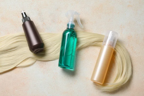 Spray bottles with thermal protection and lock of blonde hair on beige textured table, flat lay
