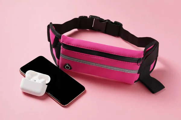 Stylish waist bag with smartphone and earphones on pink background