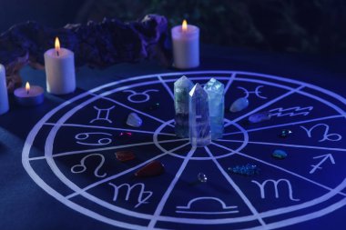 Zodyak işaretleri için doğal taşlar, astroloji çizelgesi ve koyu mavi masada yanan mumlar.