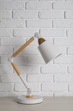 Stylish modern desk lamp on white wooden table near brick wall