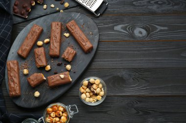 Board with delicious chocolate bars, caramel and nuts on black wooden table, flat lay. Space for text